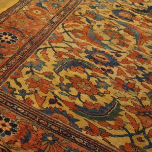 Gold and orange traditional rug with complex blue floral patterns resting on light hardwood flooring.