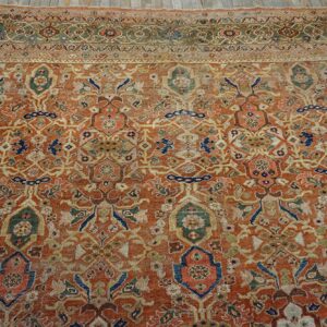 Orange-red rug with low pile and distressed traditional pattern in blue, green, and beige, placed on light wood flooring.
