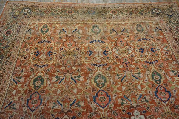 Orange-red rug with low pile and distressed traditional pattern in blue, green, and beige, placed on light wood flooring.