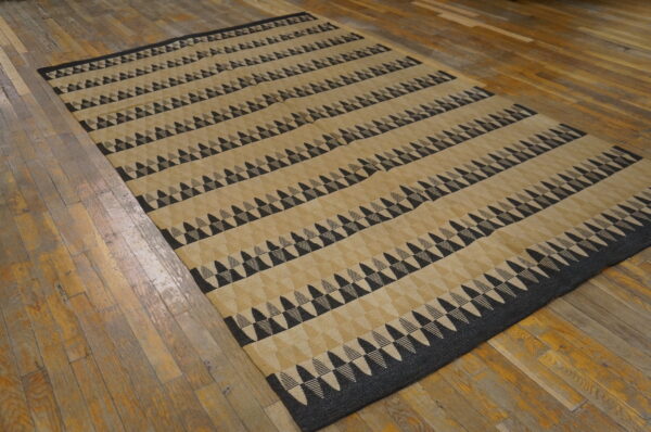 Low pile tan rug with horizontal bands of black and gray geometric triangles on worn wood planks.