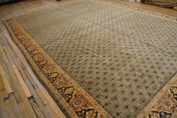 Large blue-green rug with a repeating motif pattern and wide orange and black decorative borders rests on rustic wood floors.