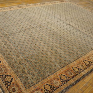 Large square rug with a dense sage and brown field pattern, framed by a wide rust and navy floral border on a wood floor.