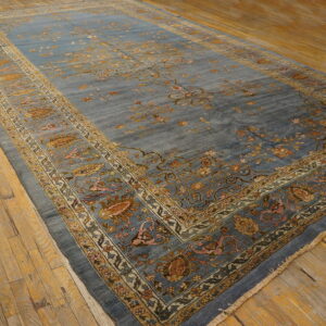 Dusty blue low-pile rug featuring traditional gold and pink floral borders rests on worn wooden planks.