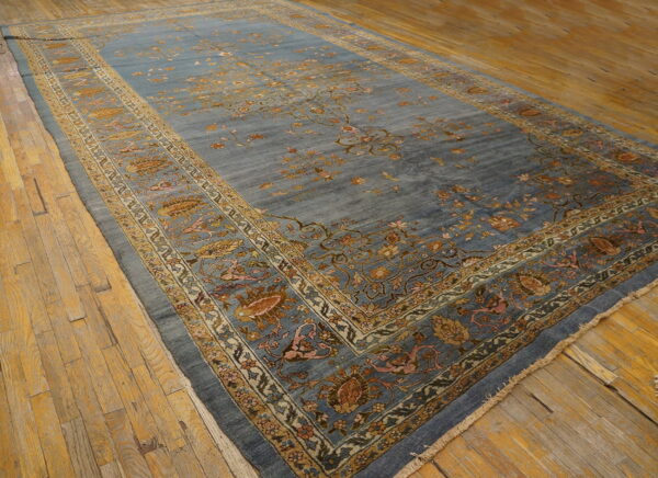 Dusty blue low-pile rug featuring traditional gold and pink floral borders rests on worn wooden planks.