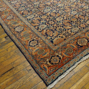 Blue and rust floral patterned rug corner with fringe displayed on worn, yellow-toned wooden floors.