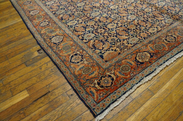 Blue and rust floral patterned rug corner with fringe displayed on worn, yellow-toned wooden floors.