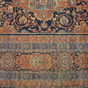 Ornate rug detail with a navy blue field, rust orange scrolling vines, and banded floral borders.