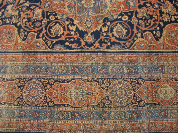 Ornate rug detail with a navy blue field, rust orange scrolling vines, and banded floral borders.