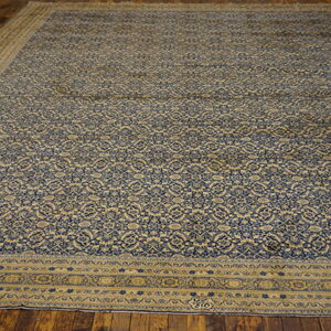 Large rug with a dense blue and tan repeating floral pattern, bordered, set on dark wood floors.