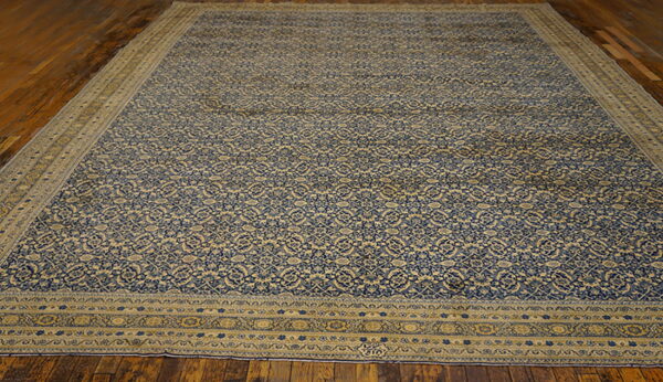 Large rug with a dense blue and tan repeating floral pattern, bordered, set on dark wood floors.