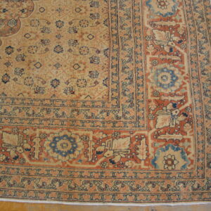 Detailed corner of a traditional rug in faded tan, rust, and blue patterns lying on wood plank flooring.