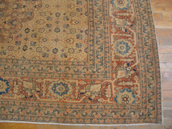 Detailed corner of a traditional rug in faded tan, rust, and blue patterns lying on wood plank flooring.