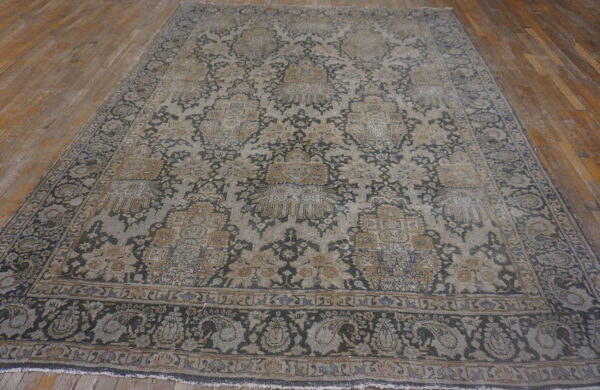 A large, low-pile rug with complex floral motifs in charcoal, gray, and tan sits on wooden planks.