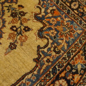 Muted gold rug with traditional dark blue and rust floral patterns rests on distressed wooden floors.