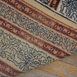 Beige rug with striped borders of navy and rust geometric patterns rests on distressed wooden floorboards.