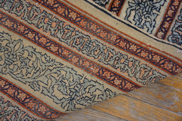 Beige rug with striped borders of navy and rust geometric patterns rests on distressed wooden floorboards.