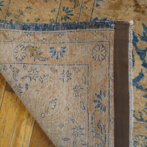 Corner of a low-pile tan and blue distressed rug folded back, exposing its woven backing on hardwood.