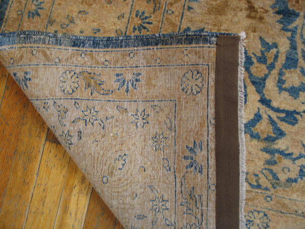 Corner of a low-pile tan and blue distressed rug folded back, exposing its woven backing on hardwood.
