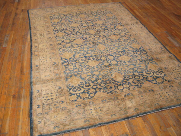 Low-pile blue and tan traditional rug with ornate pattern covering wide plank wood flooring.