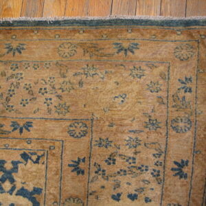Corner of a golden rug featuring intricate blue floral patterns, a dark border, and wood plank flooring.