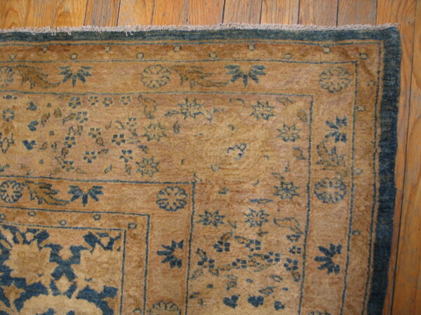 Corner of a golden rug featuring intricate blue floral patterns, a dark border, and wood plank flooring.