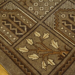 Detail of a brown geometric rug featuring checkered and zigzag borders and a stylized tree motif on hardwood.