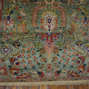 Textured green rug with red borders and intricate floral and avian patterns rests on rustic wood flooring.