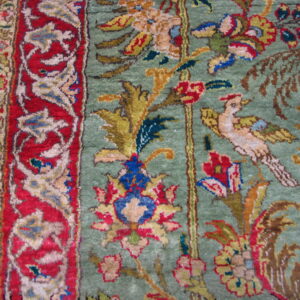 Close-up shows a multicolored traditional rug, green field, red border, and floral and bird pattern on wood.