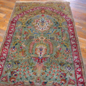 Green and red pictorial rug featuring a mihrab design and dense floral motifs on hardwood planks.