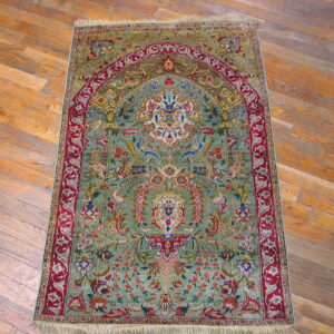 Ornate low-pile rug with green and red floral mihrab pattern displayed on light wood floor.