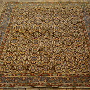 Traditional low pile rug in ochre, rust, and blue geometric floral patterns on distressed wood flooring.