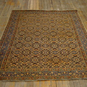 A low-pile rug featuring complex repeating patterns in tan, blue, and rust rests on distressed wooden floors.