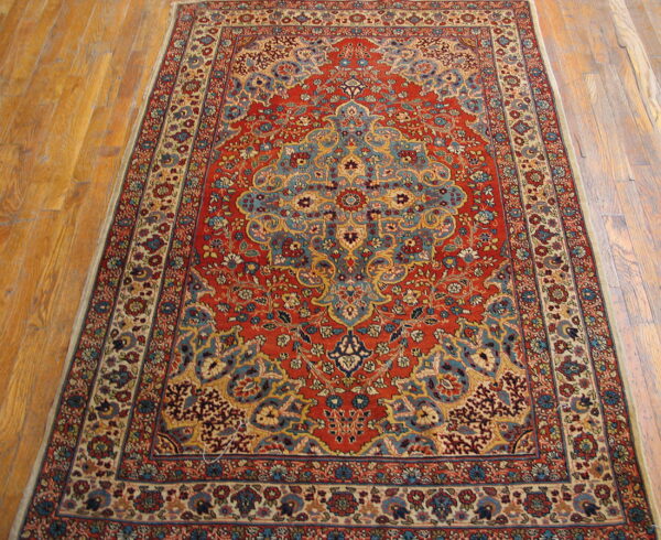 A richly patterned low-pile rug with a red field and blue floral motifs on medium-toned wood flooring.