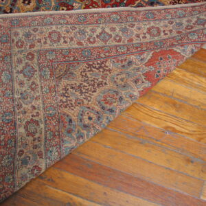 Red and blue traditional rug is folded over, exposing its woven structure on light brown hardwood flooring.