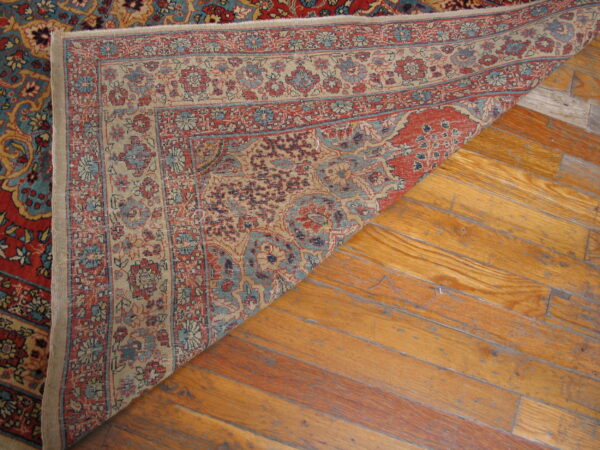 Red and blue traditional rug is folded over, exposing its woven structure on light brown hardwood flooring.
