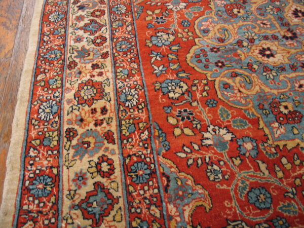 Ornate floral rug, featuring a rust red field, teal accents, and a wide cream border strip on wooden floorboards.