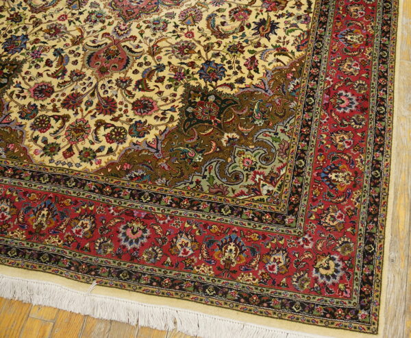 Intricate cream rug corner featuring a dense floral pattern and a wide red border on light wood flooring.
