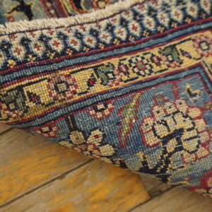 Intricate blue, gold, and red patterned rug edge rolled over distressed yellow wood flooring.