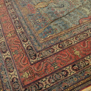 Ornate low-pile rug featuring a blue field with tree pattern and wide red floral borders on hardwood.