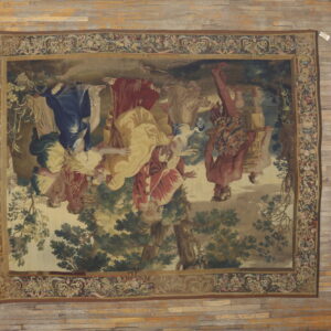 Rectangular tapestry rug with blue, red, and yellow figures in a landscape border, displayed on wide wood plank flooring.