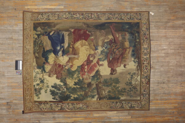 Rectangular tapestry rug with blue, red, and yellow figures in a landscape border, displayed on wide wood plank flooring.