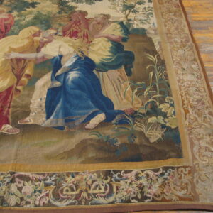 Figural tapestry rug in blue, red, and green features decorative floral borders lying on wide plank wood floors.