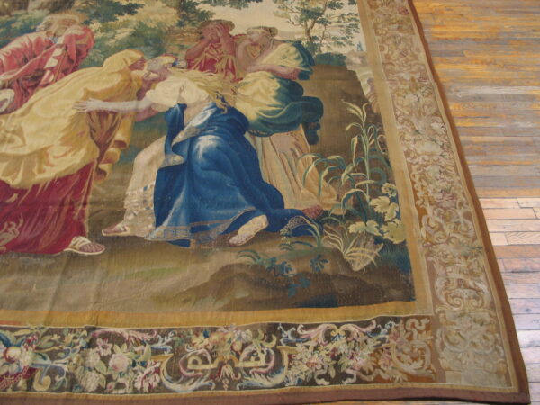 Figural tapestry rug in blue, red, and green features decorative floral borders lying on wide plank wood floors.