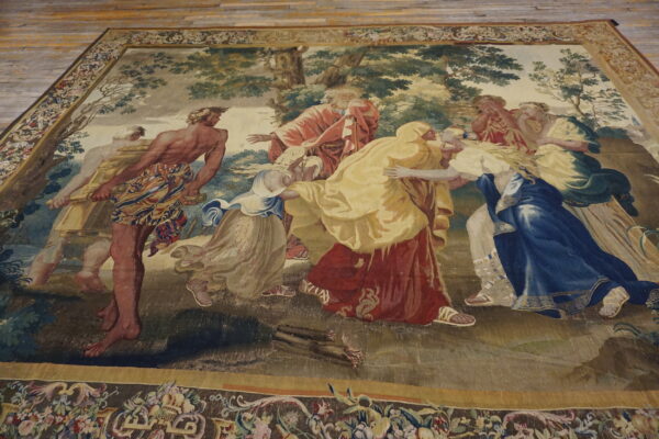 Low-pile rug displays a classical figurative scene in blue, red, and gold, bordered by florals, on wood.