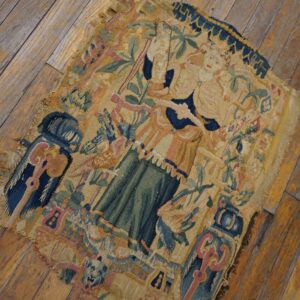 A worn flat-weave fragment with figural and foliage patterns in tan, blue, green, and pink rests on wood flooring.