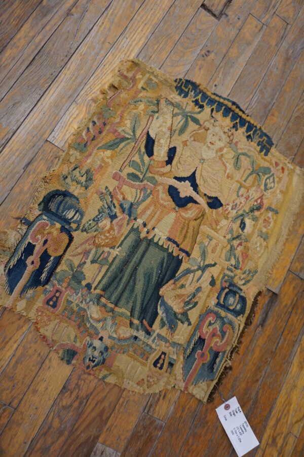 A worn flat-weave fragment with figural and foliage patterns in tan, blue, green, and pink rests on wood flooring.