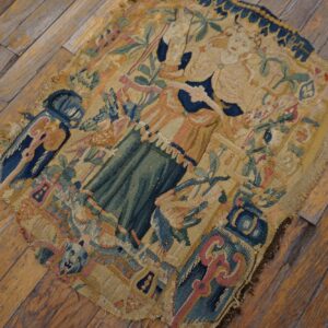 Frayed flatweave textile fragment showing blue, green, and tan figures and florals on distressed wood flooring.