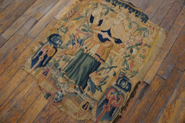 Frayed flatweave textile fragment showing blue, green, and tan figures and florals on distressed wood flooring.