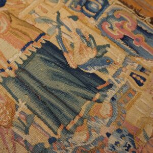 Detailed view of a tapestry-like rug fragment in faded blues, greens, and pinks lying on hardwood flooring.