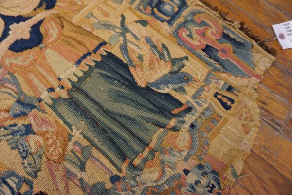 Detailed view of a tapestry-like rug fragment in faded blues, greens, and pinks lying on hardwood flooring.